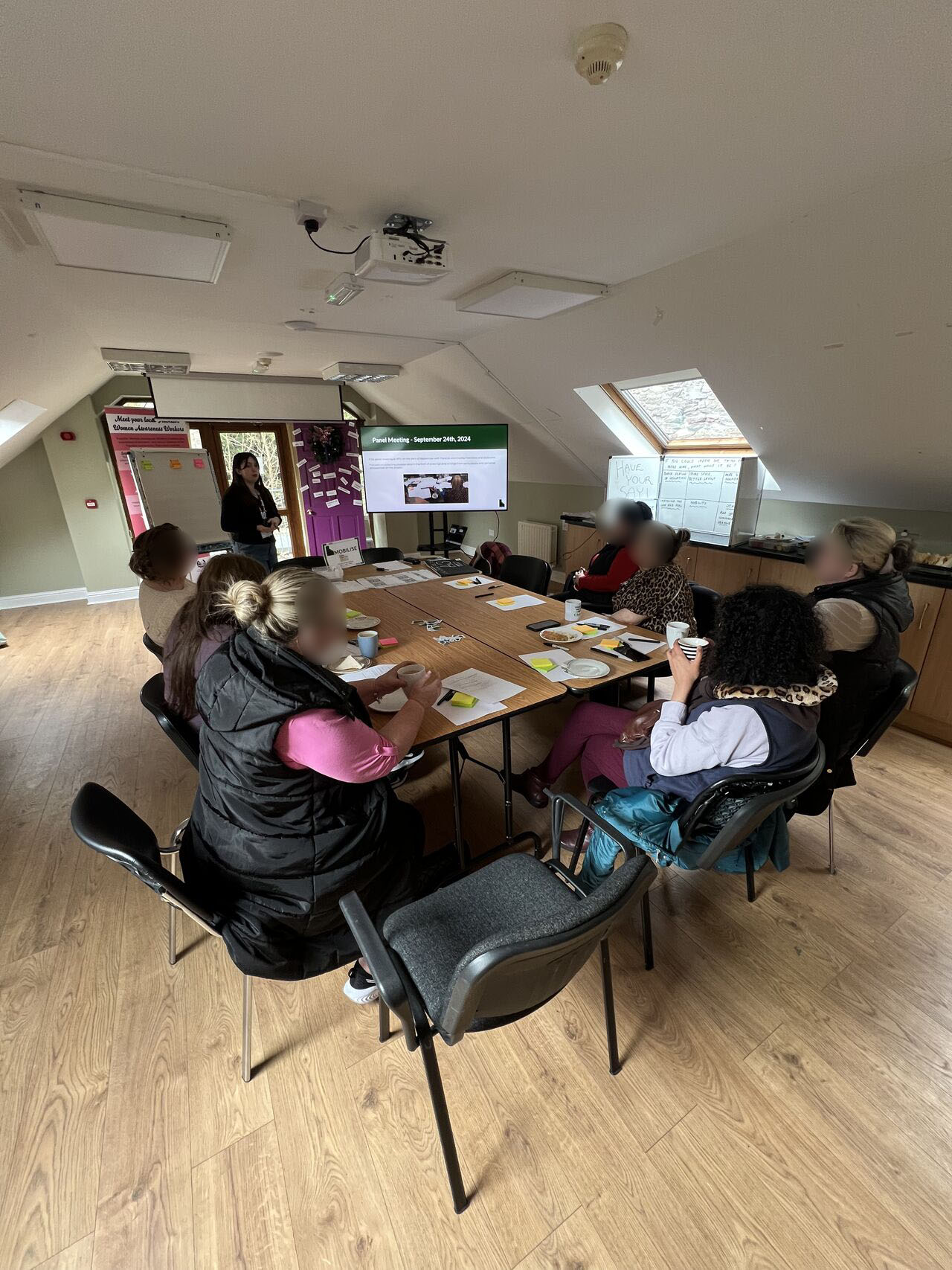 An example of a previous Irish Traveller community workshop. Participants are pictured gathered around a table with drawing materials, watching a researcher present on a large digital whiteboard.